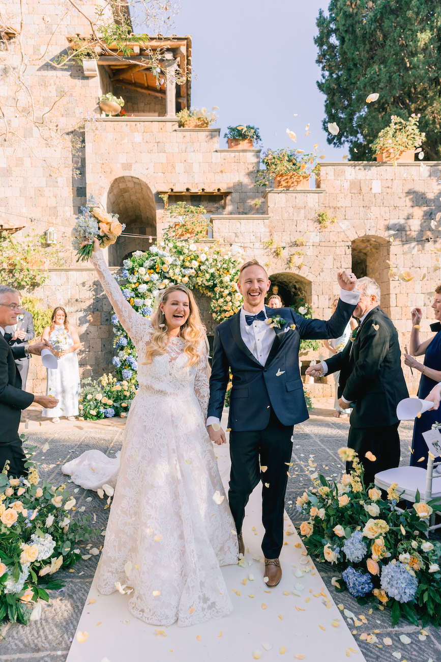 A bride and groom walk hand in hand down an aisle outdoors, smiling and raising their arms as flower petals fall around them, surrounded by guests and floral decorations.
