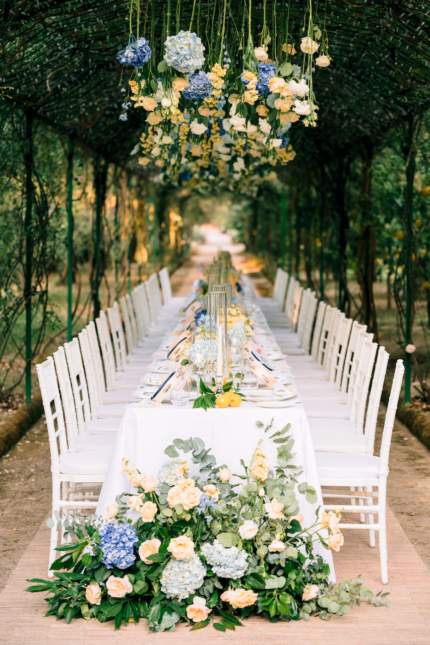 A long outdoor dining table with white chairs is decorated with white and blue flowers, greenery, and hanging floral arrangements under an arched trellis.