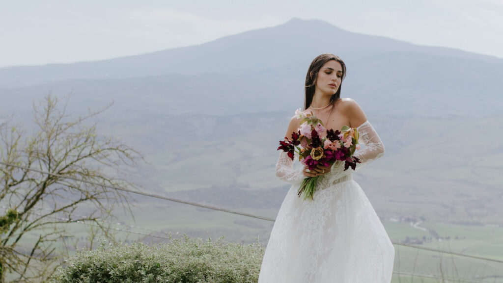 A woman in a white wedding dress holds a bouquet of flowers, standing outdoors with mountains and greenery in the background.