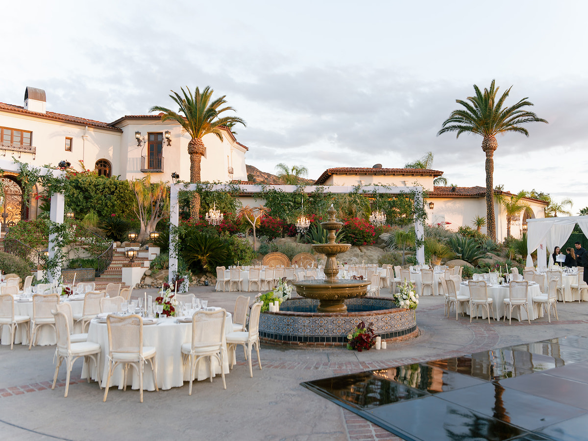 Outdoor courtyard set for an event with round tables, white chairs, a central fountain, palm trees, and a large Mediterranean-style building in the background.