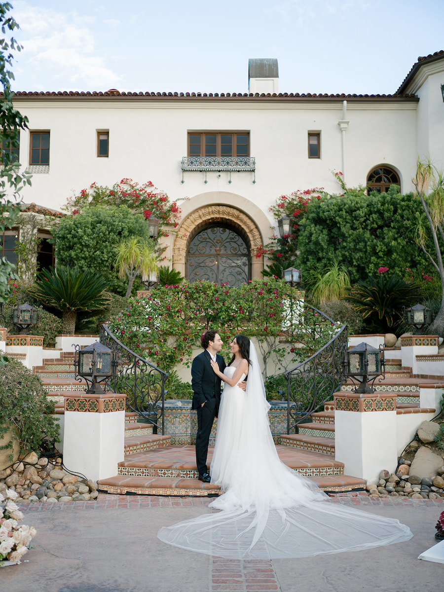 A bride and groom stand facing each other on tiled steps outside a large, white Mediterranean-style building with lush greenery and flowers.