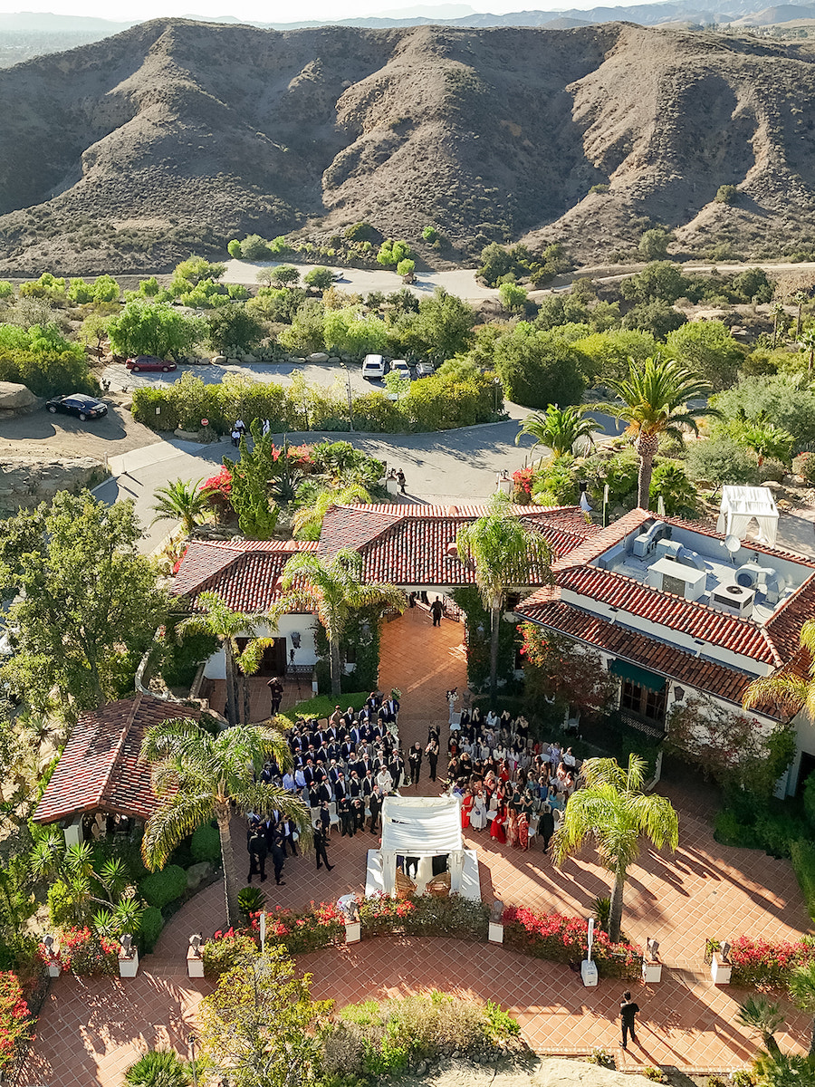 Aerial view of an outdoor wedding ceremony at a villa with red-tiled roofs, surrounded by trees and mountains in the background.