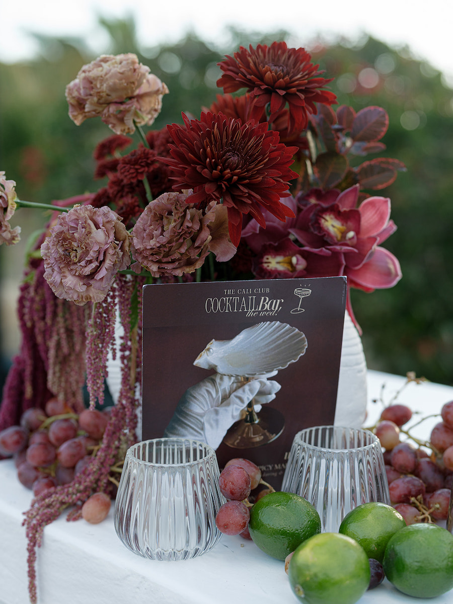 A floral arrangement with red and pink flowers, grapes, limes, two glass candleholders, and a cocktail menu on a white tablecloth.