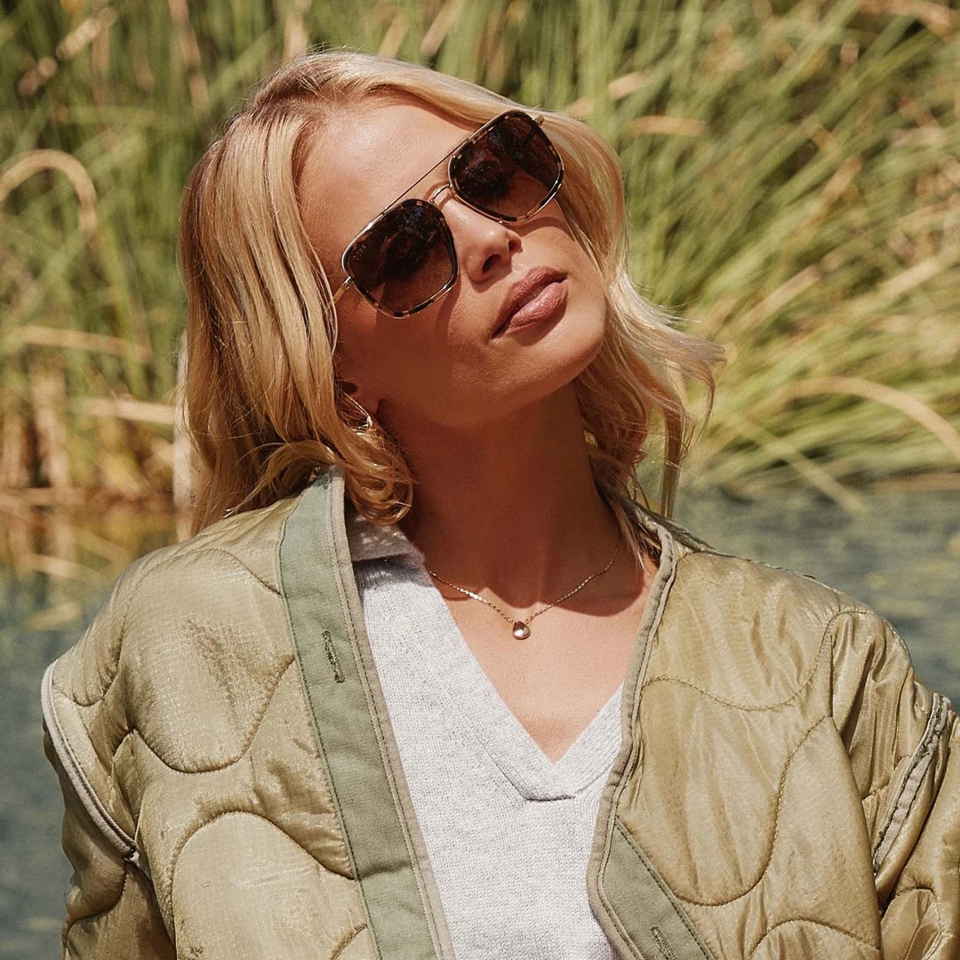 Woman with blonde hair wearing sunglasses, a light jacket, and a necklace stands outdoors in front of tall green grass, facing the camera with a slight head tilt.
