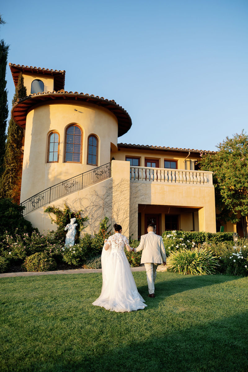 A bride and groom walk on the lawn toward a large Mediterranean-style villa under a clear sky on their Paso Robles wedding day.