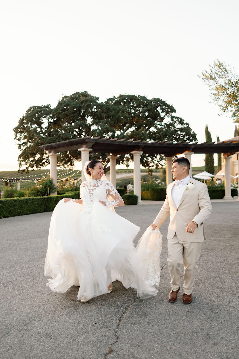 A bride in a white gown and a groom in a beige suit smile at each other while walking outdoors near large trees and a pergola.