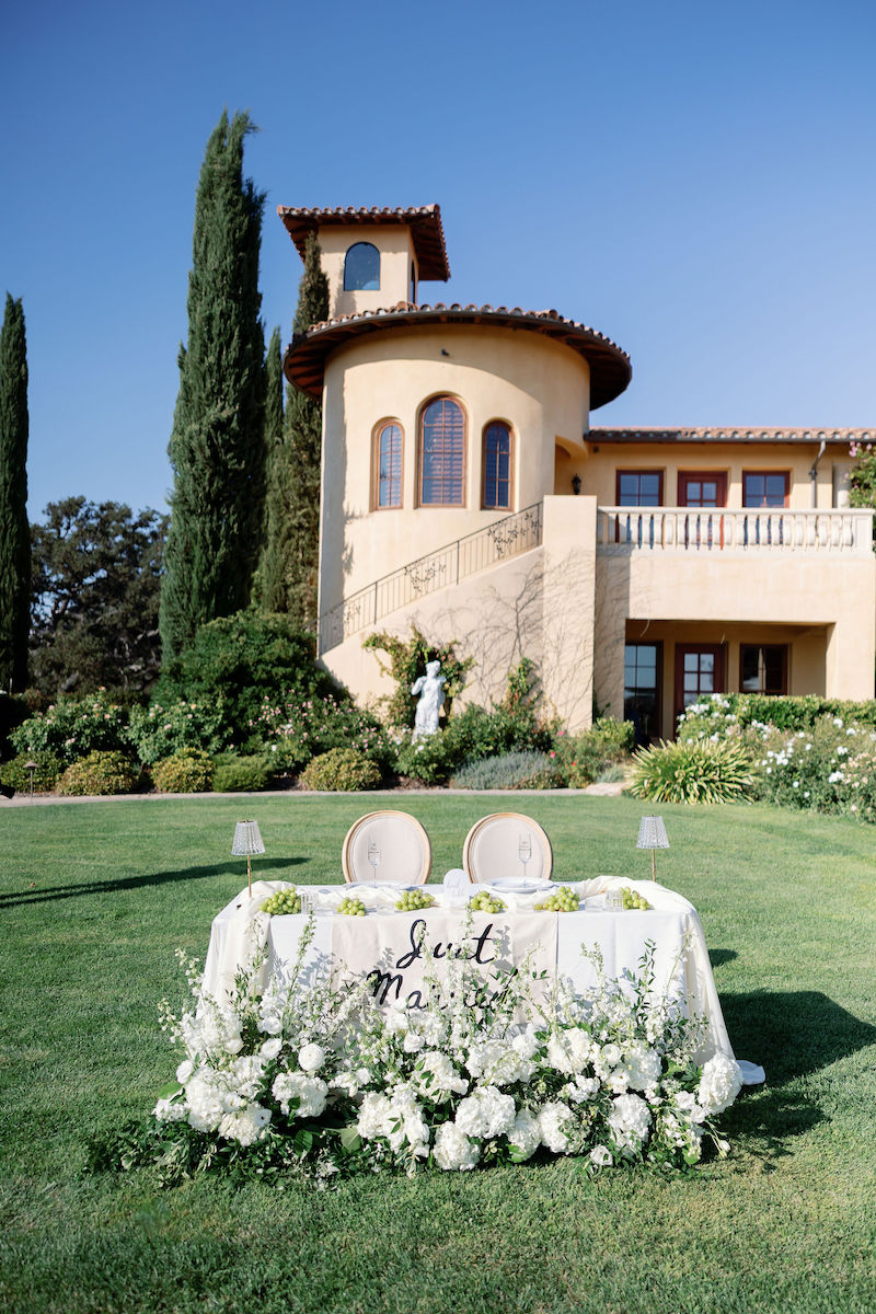 A decorated sweetheart table with white flowers on a lawn, in front of Villa San Juliette with tall cypress trees and blue sky.