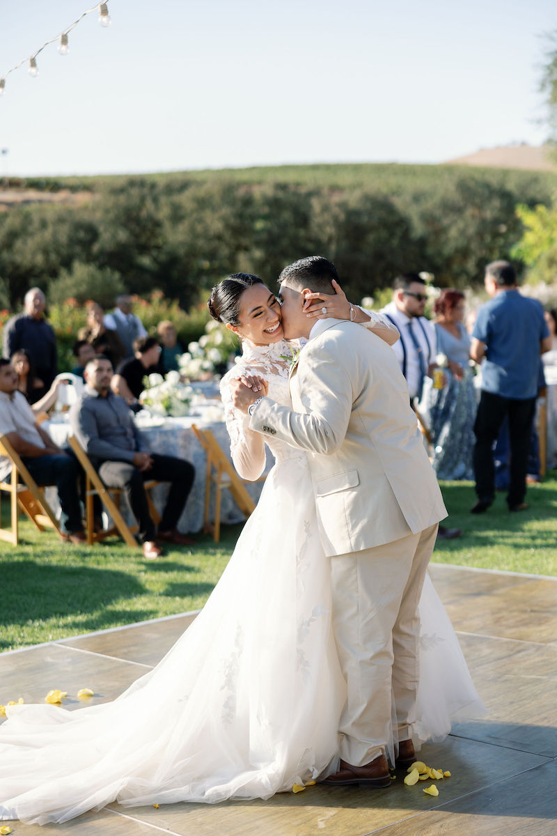 A bride and groom dance together outdoors at their wedding reception, with guests seated at tables in the background.