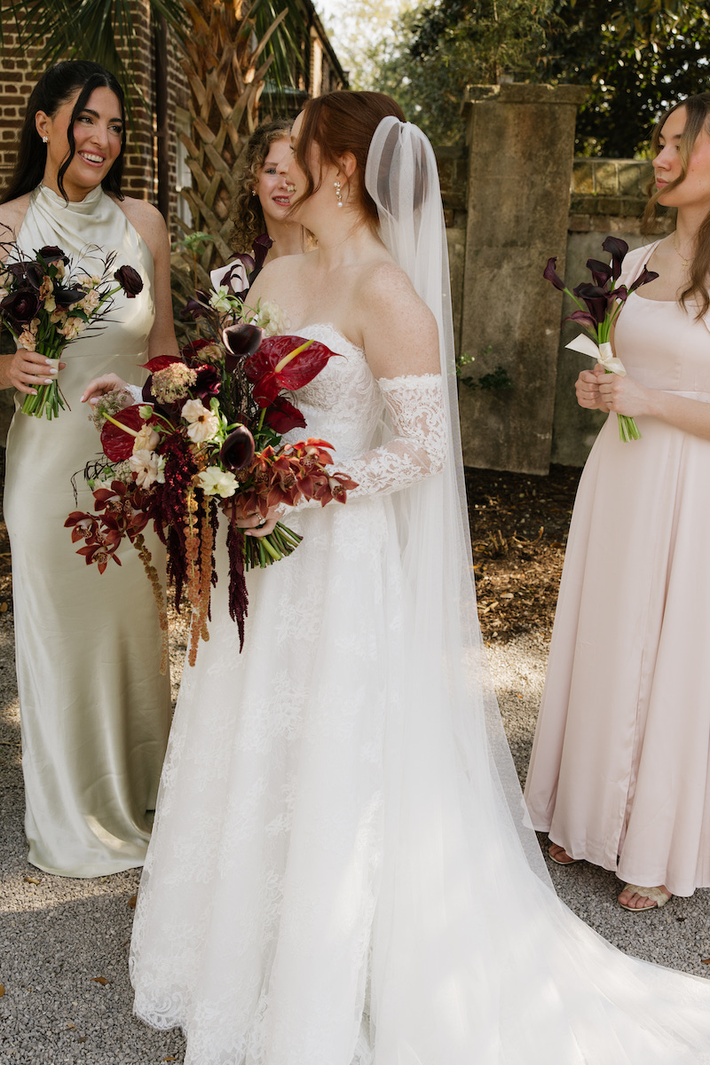 A bride in a white dress and veil stands outdoors holding a bouquet, surrounded by three bridesmaids in light-colored dresses, all smiling and talking.