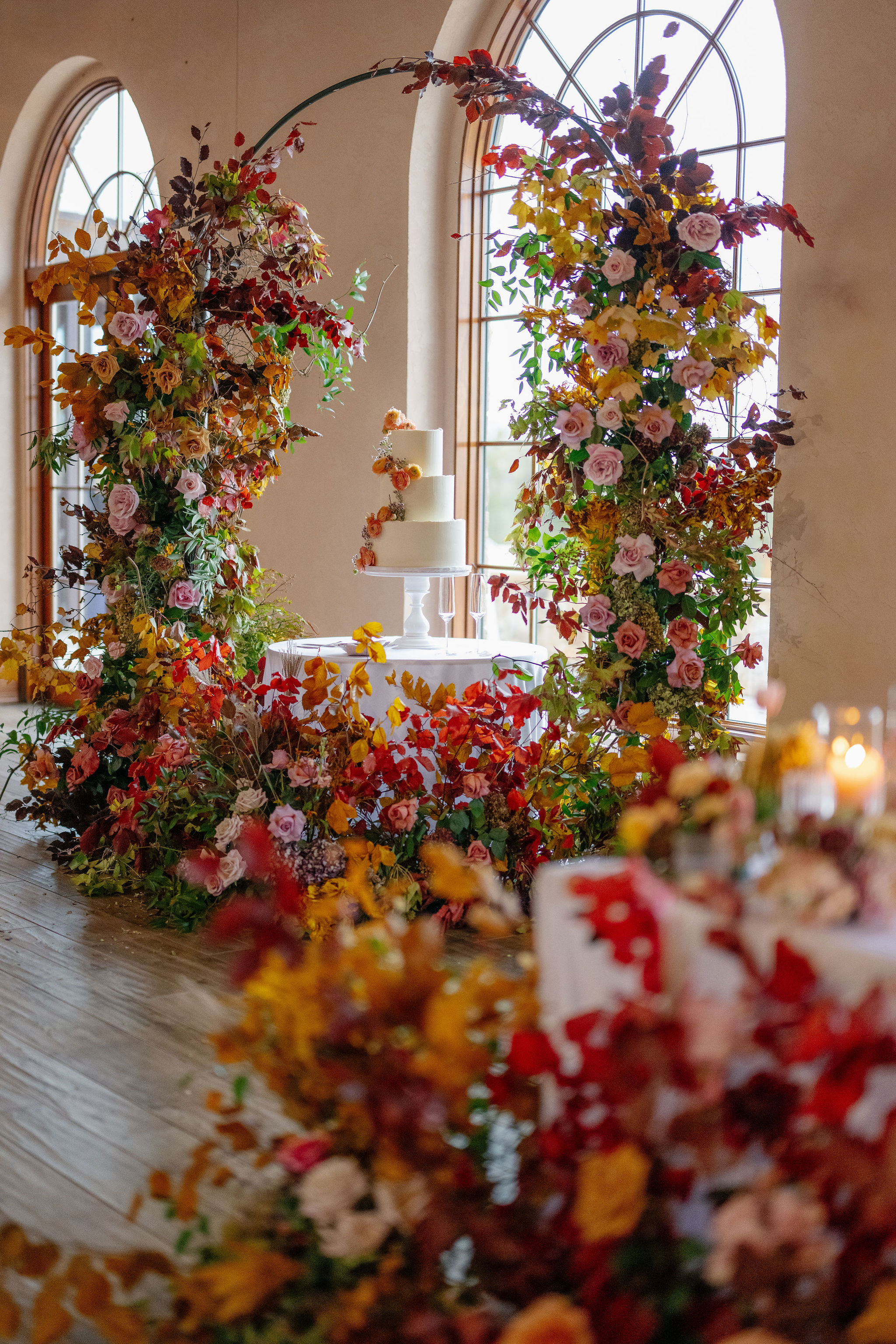 A wedding cake stands on a round table framed by floral arches with colorful flowers and greenery, set in front of tall arched windows.
