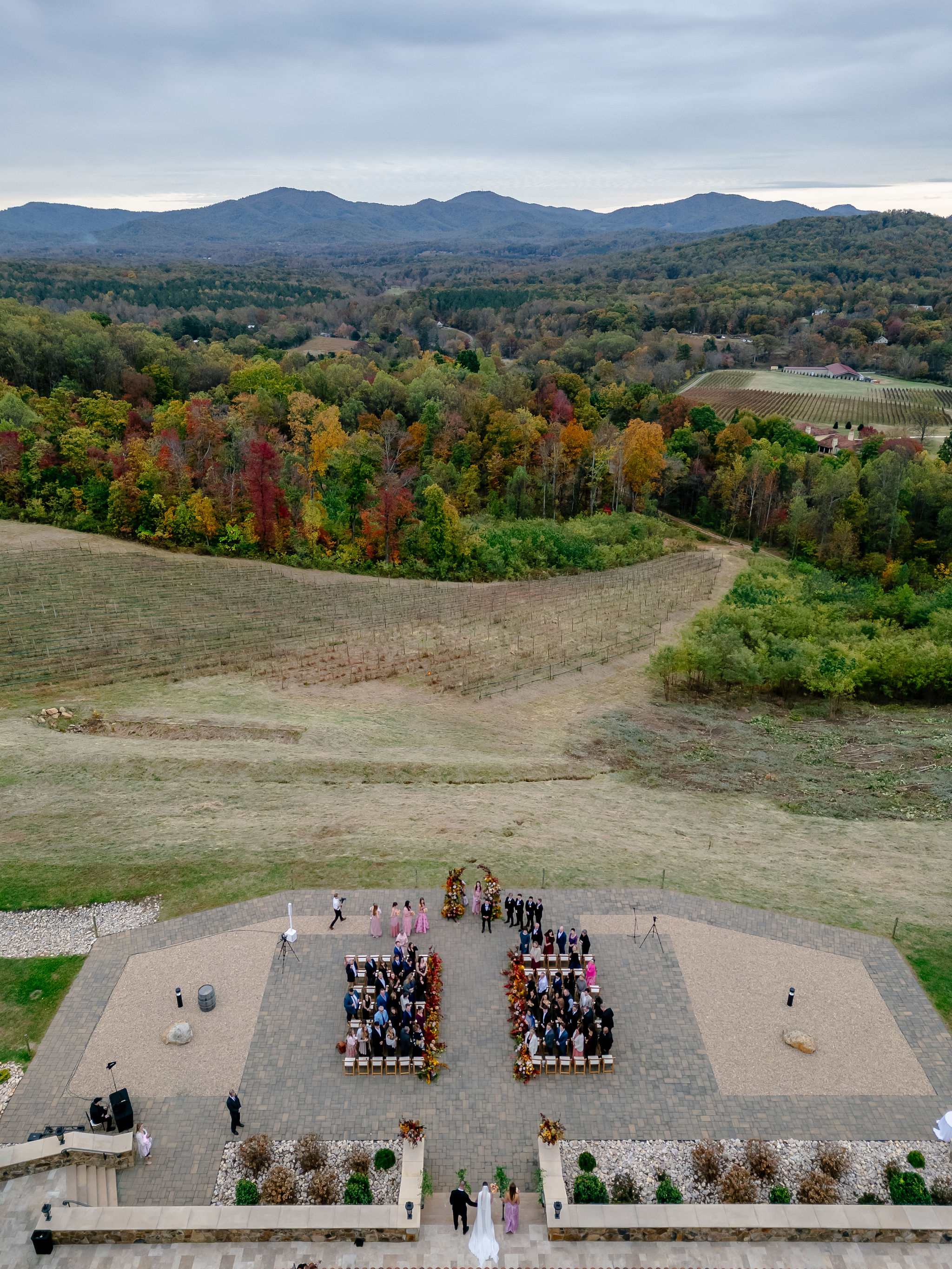 Outdoor wedding ceremony with guests seated in rows on a paved area, surrounded by natural landscape and distant mountains under a cloudy sky.
