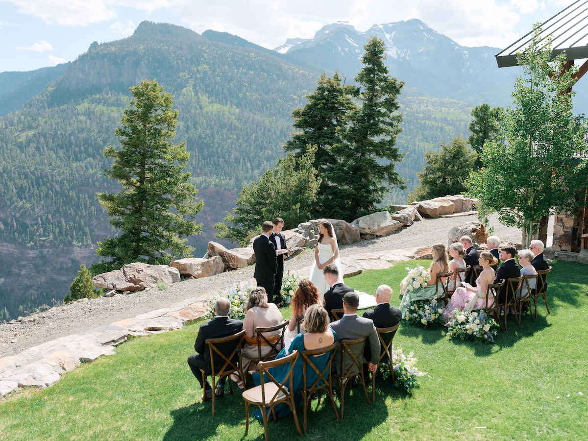 An outdoor wedding ceremony with guests seated on a lawn, set against a backdrop of mountains and trees under a clear sky.