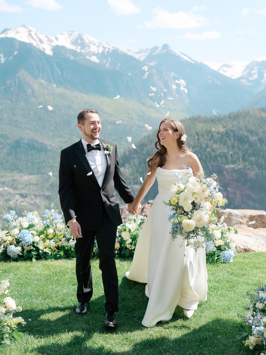 A bride and groom walk hand in hand outdoors, smiling, with mountains and blue sky in the background and flowers on the grass around them.