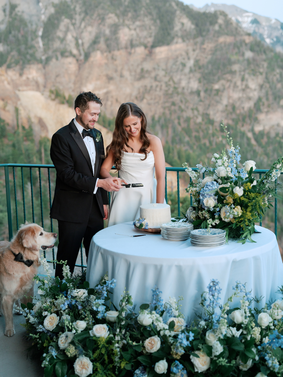 A couple in formal attire cuts a small cake on an outdoor table decorated with blue and white flowers, with a golden retriever beside them and mountains in the background.