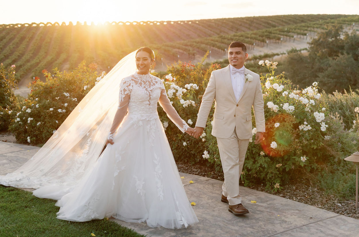 Bride and groom walk hand in hand outside at sunset, with vineyards and white flowers in the background.