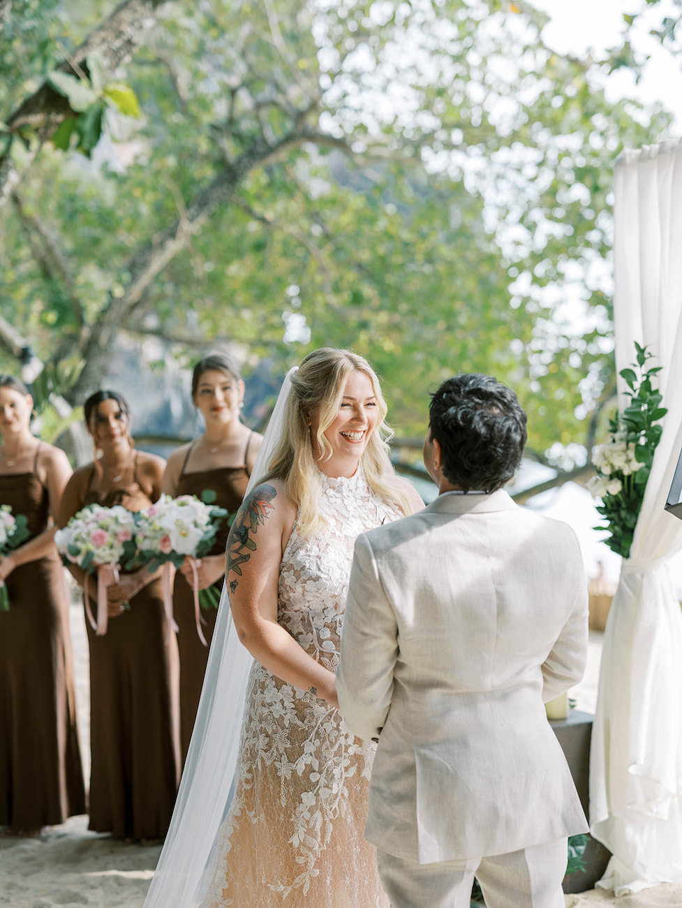 A bride and groom stand facing each other during an outdoor wedding ceremony, with bridesmaids in brown dresses holding bouquets in the background.
