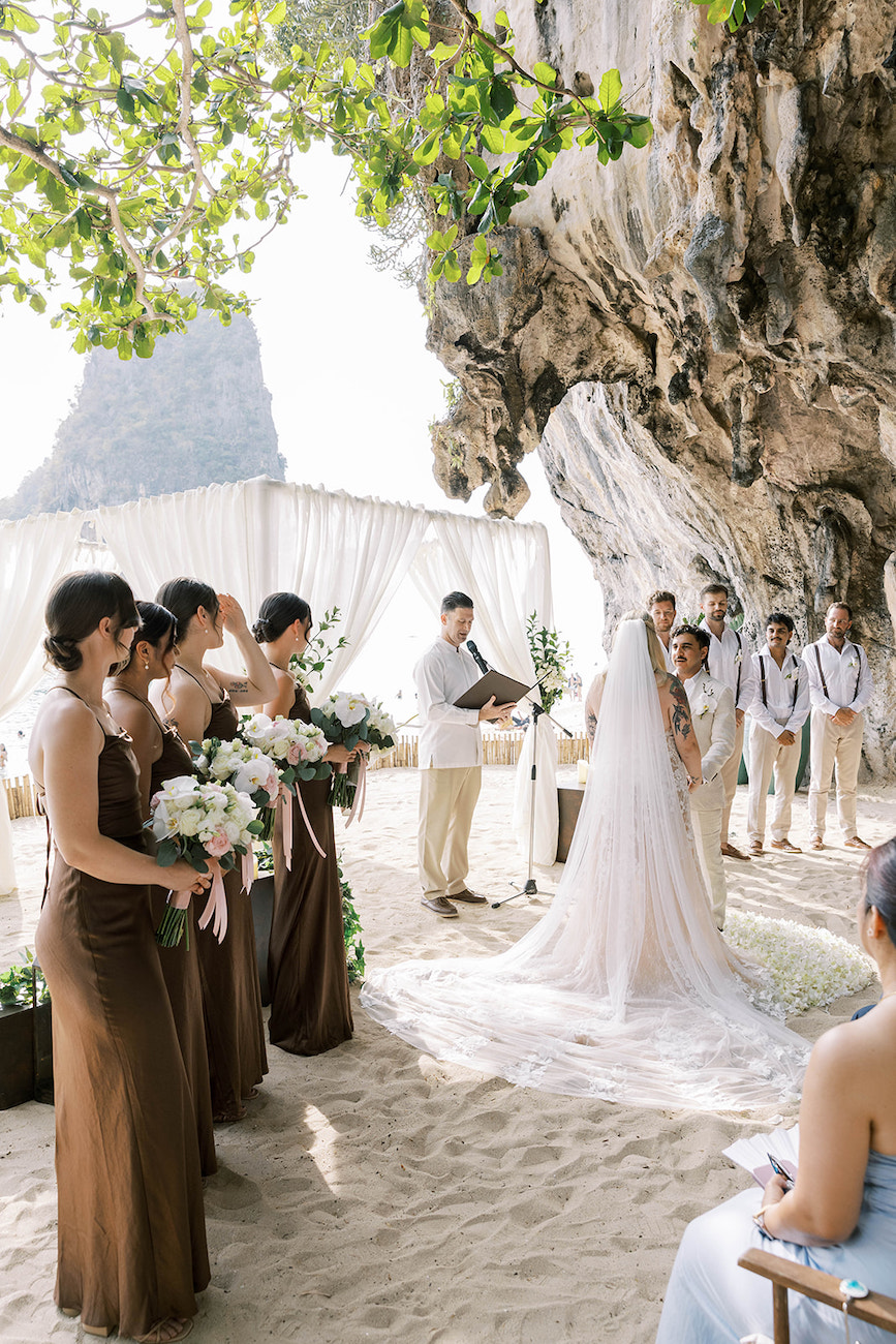 A bride and groom stand under a rocky cliff during an outdoor beach wedding ceremony, with bridesmaids in brown dresses holding bouquets.