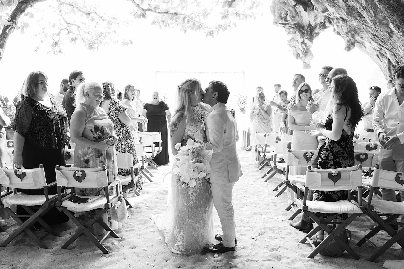 A couple stands and kisses at the end of a sandy aisle, surrounded by guests seated on folding chairs at a wedding ceremony under a canopy of trees.