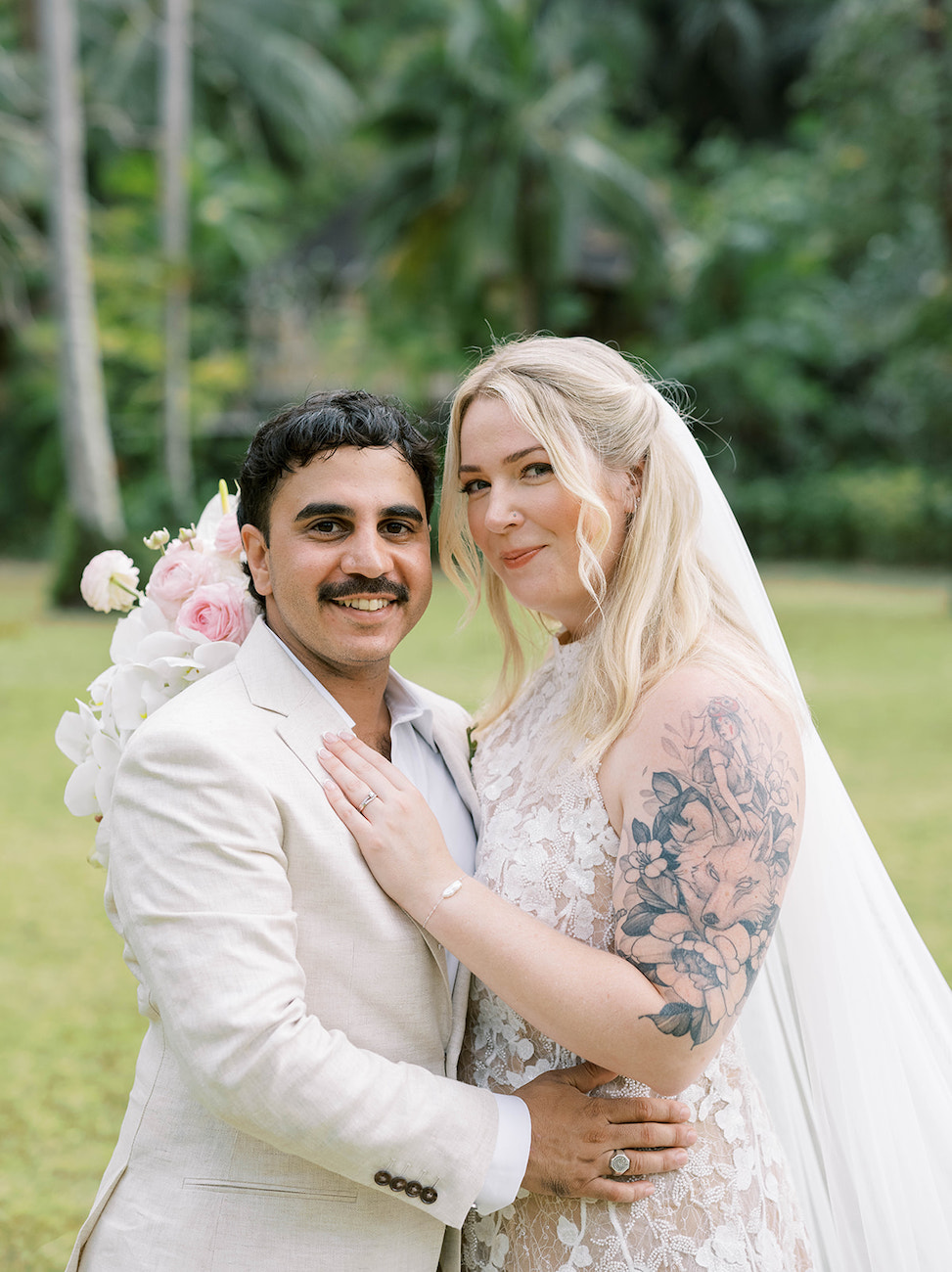 A couple in wedding attire poses outdoors; the groom wears a light suit and the bride wears a lace dress with a floral tattoo and holds a bouquet.