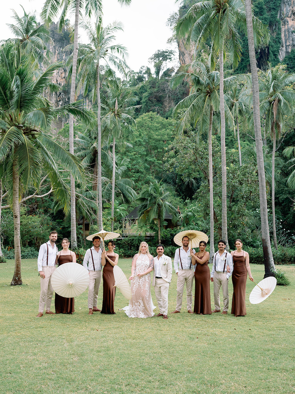 A wedding party poses outdoors in tropical surroundings, with bridesmaids in brown dresses and groomsmen in beige suits holding large white parasols.