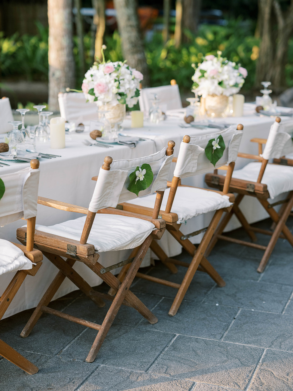 Outdoor table set with white linens, wooden chairs with white covers, and floral centerpieces in vases; greenery visible in the background.