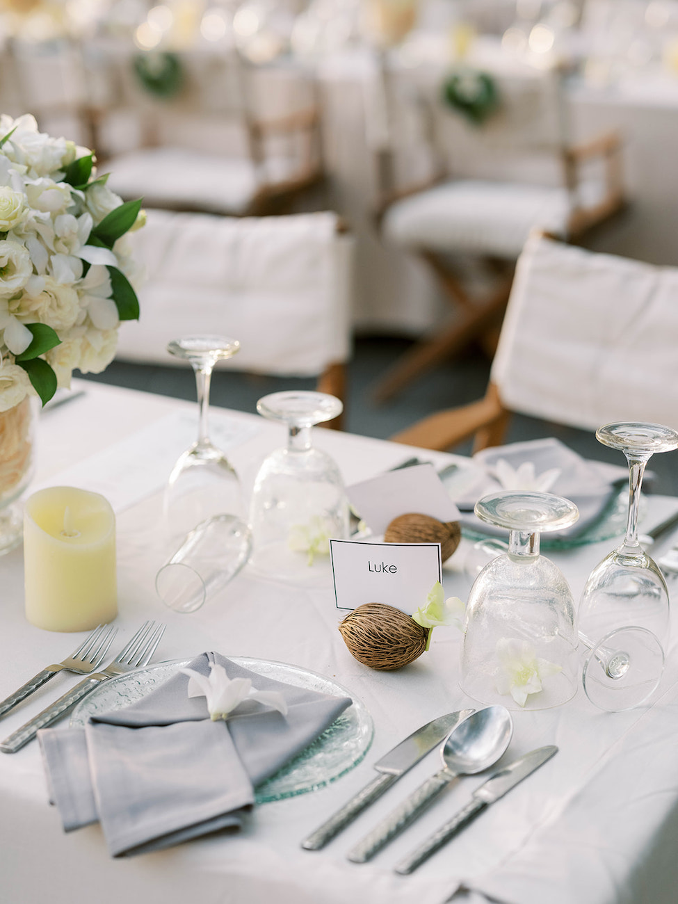 Elegant table setting with white linens, glassware, silver cutlery, a candle, white flowers, and a place card labeled “Luke” next to a coconut, set for a formal event or dinner.