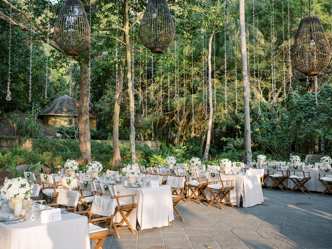 Outdoor event with multiple tables covered in white linens and floral centerpieces, surrounded by wooden chairs and hanging wicker lanterns, set in a lush, green forest environment.