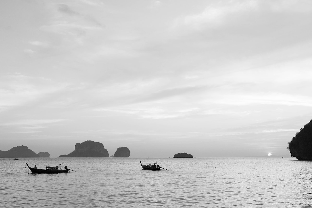 Two small boats float on calm water at sunset, with distant rocky islands and a cloudy sky in the background.