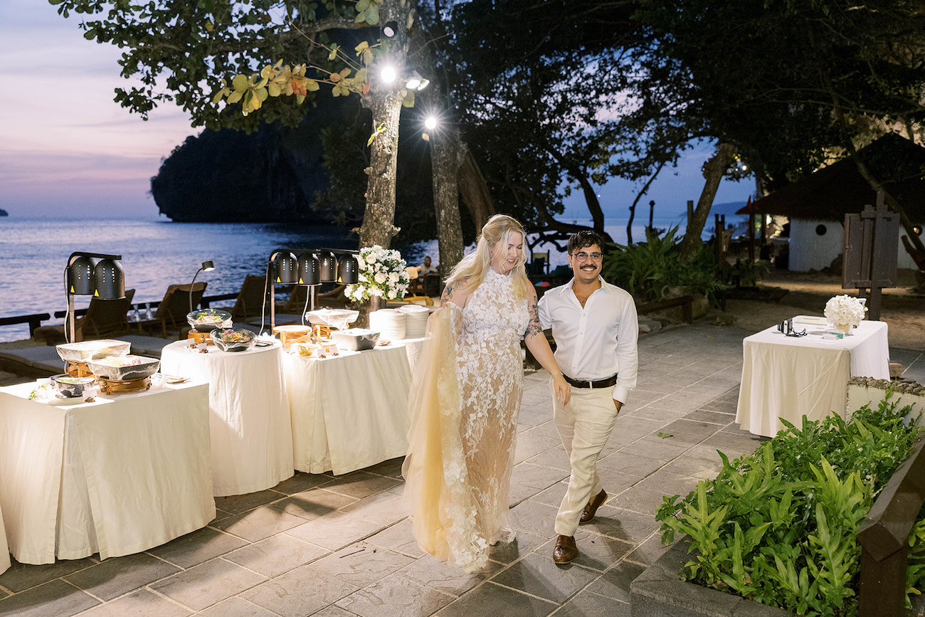 A couple walks hand in hand at an outdoor evening event near the sea, with decorated buffet tables and trees around them.