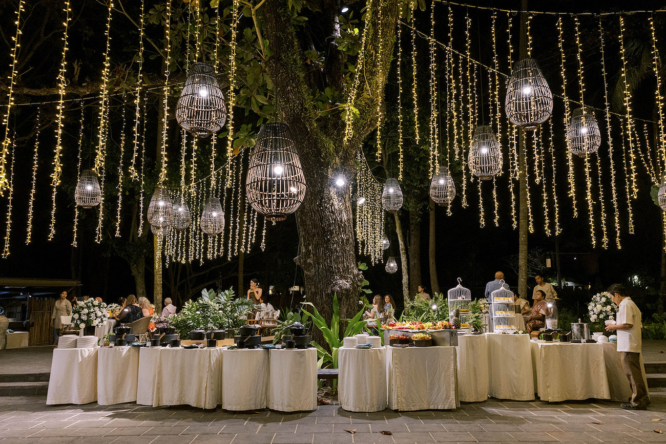 Outdoor evening event with round tables, food displays, and people mingling under hanging lights and lanterns in a tree-filled setting.