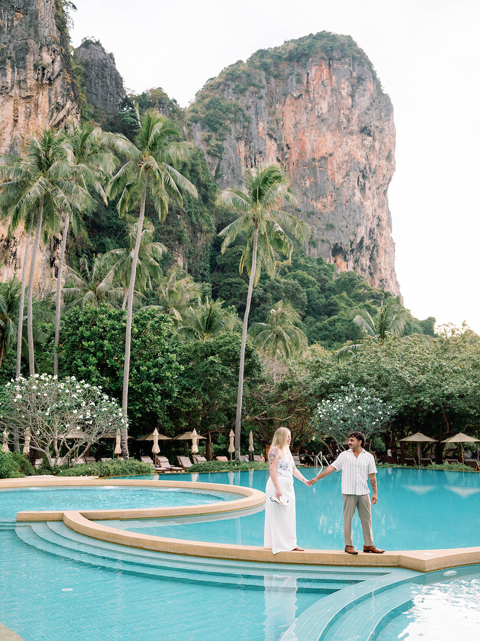 A couple stands hand in hand by a curved pool, surrounded by palm trees and lush greenery, with a large rocky cliff in the background.