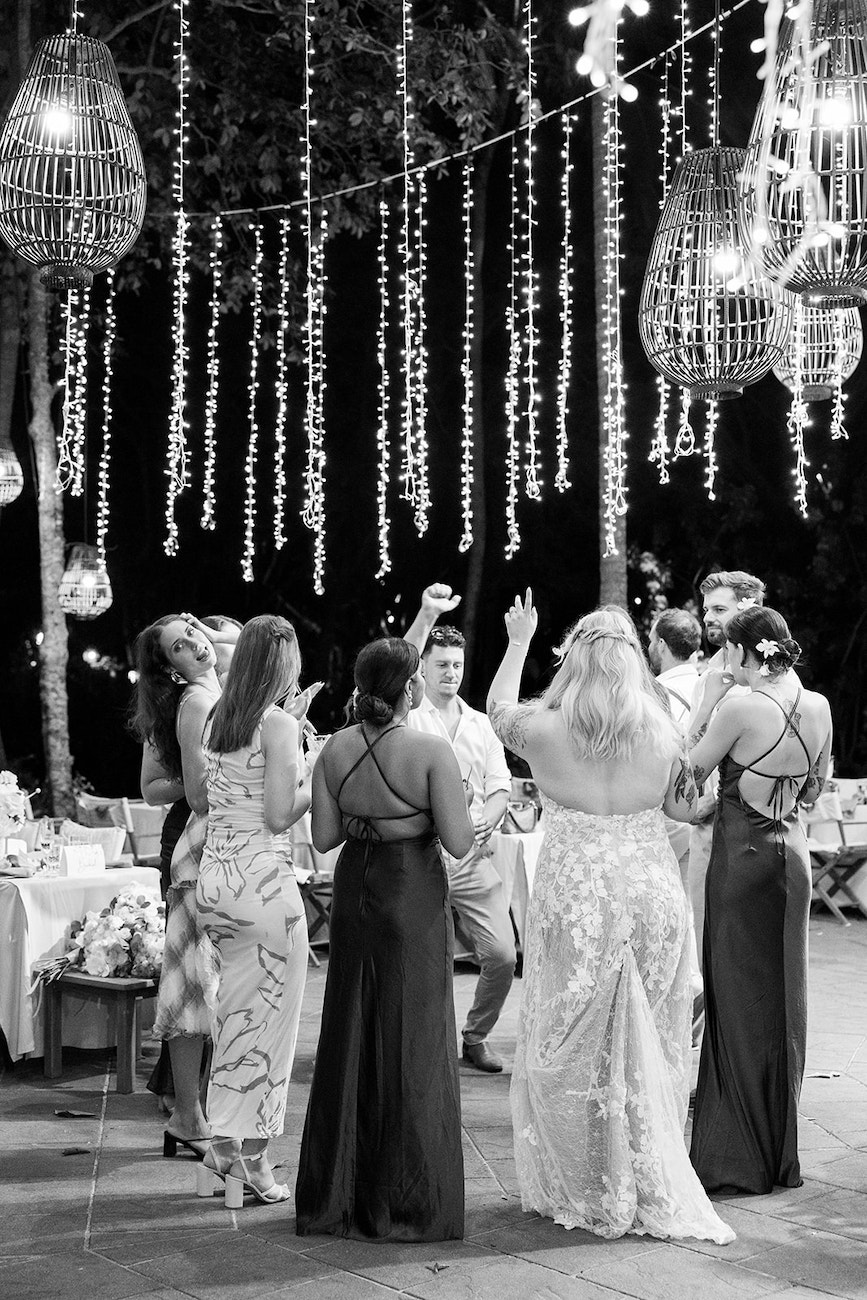 A group of people in formal attire dance together outdoors at night under hanging string lights and lanterns.