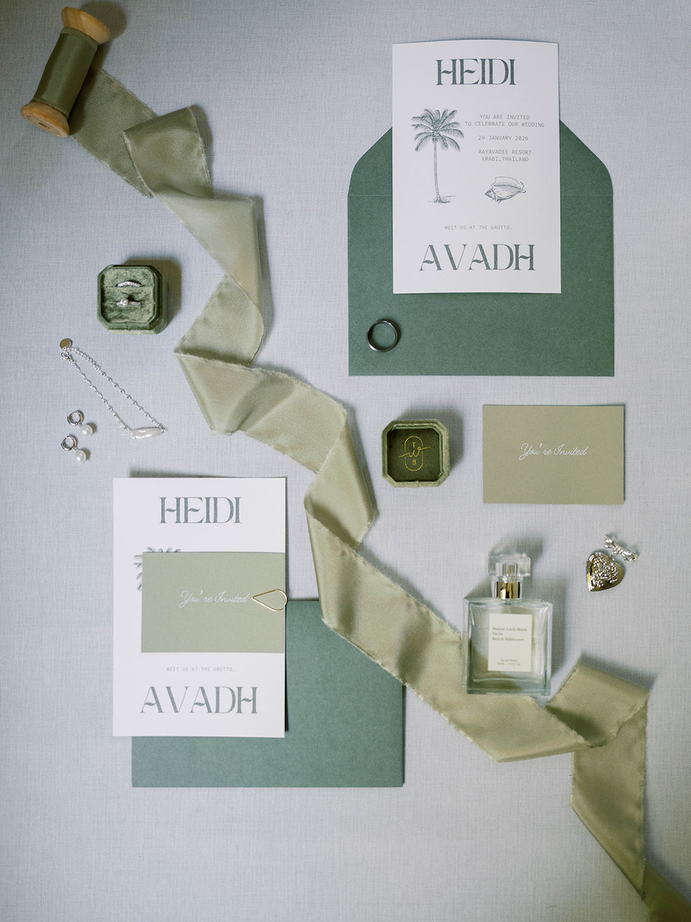 Flat lay of wedding invitation suite with green envelopes, ribbon, perfume bottle, rings, jewelry, and two hexagonal boxes on a light background.