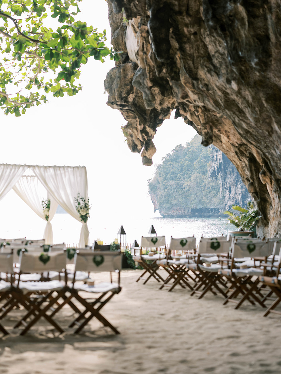 Rows of chairs face a simple wedding arch on a sandy beach, set beneath a large rocky cliff with greenery and ocean in the background.