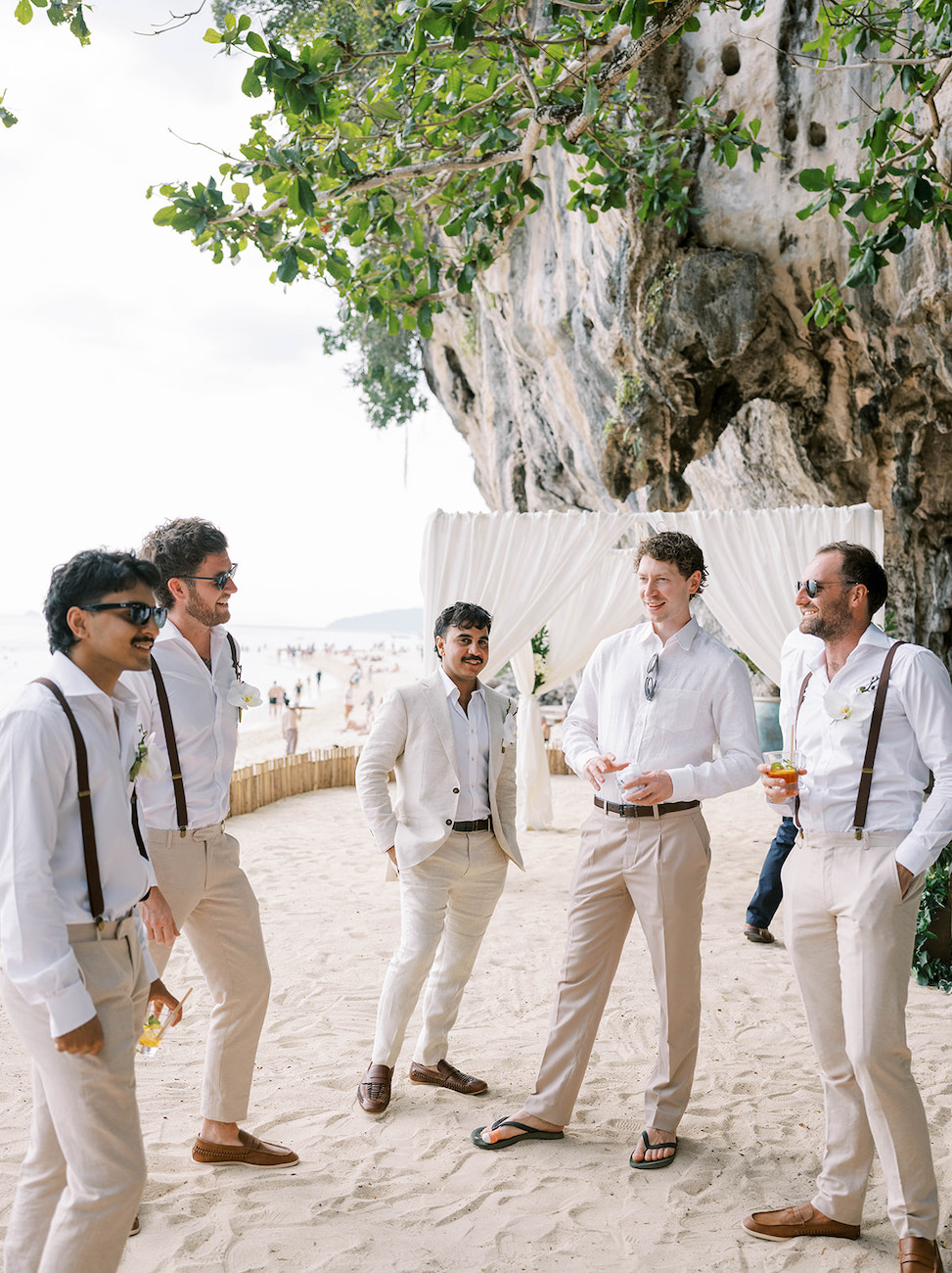 Five men in white shirts and beige pants stand and talk on a sandy beach near a large rock, with trees and beach decorations in the background.