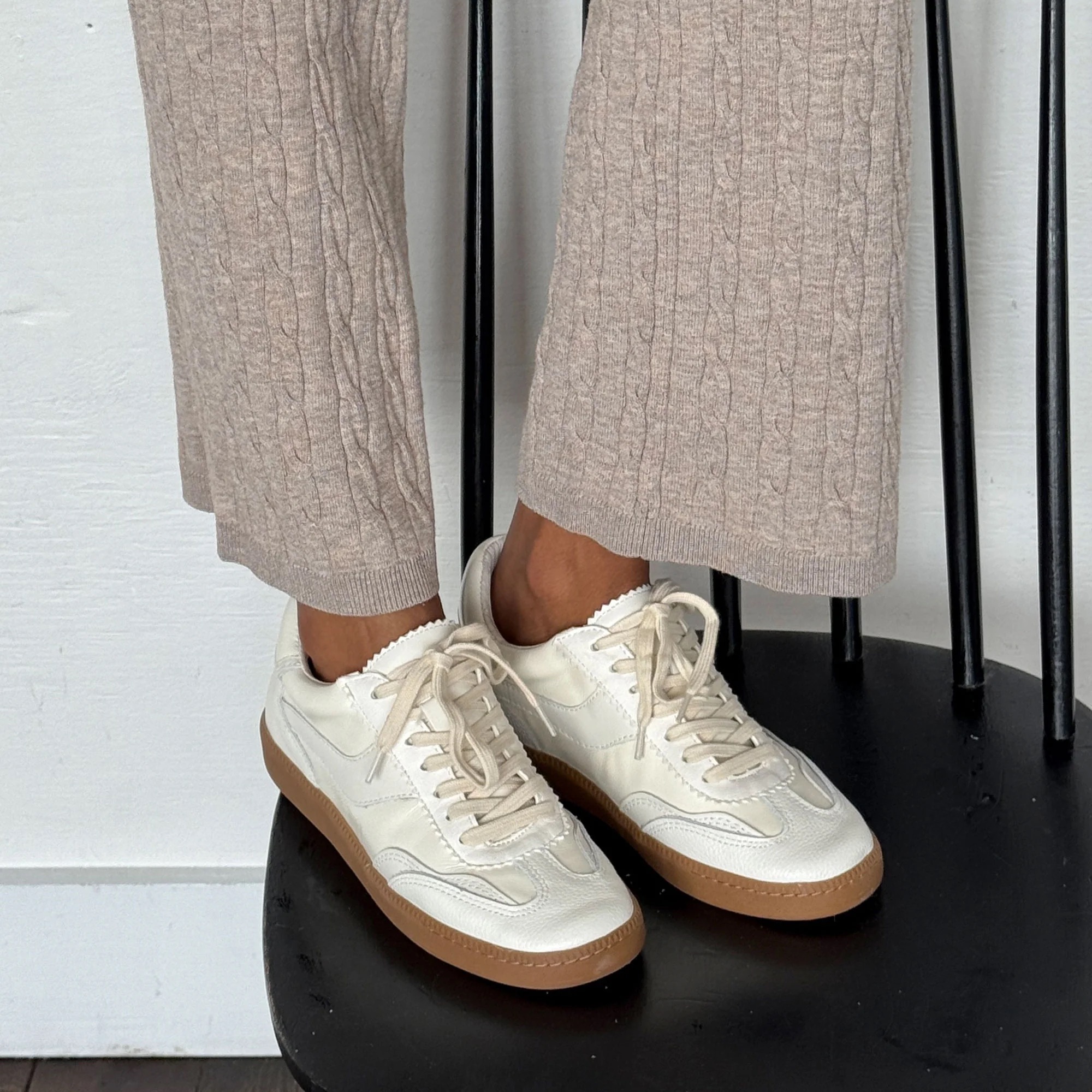 Person wearing beige textured pants and white sneakers with gum soles, standing on a black surface against a white background.