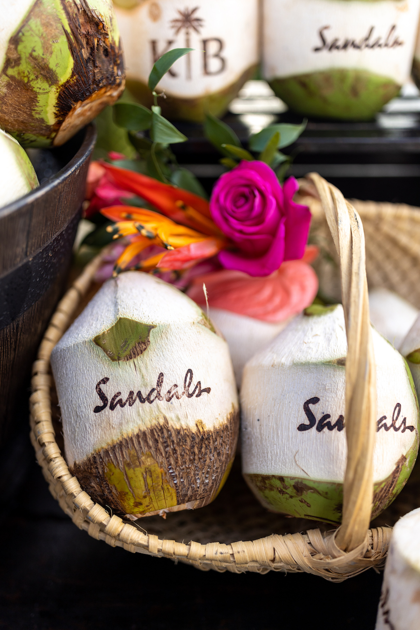 Basket with coconuts labeled “Sandals,” a pink rose, and tropical flowers. A bowl with more coconuts is partially visible on the left side.