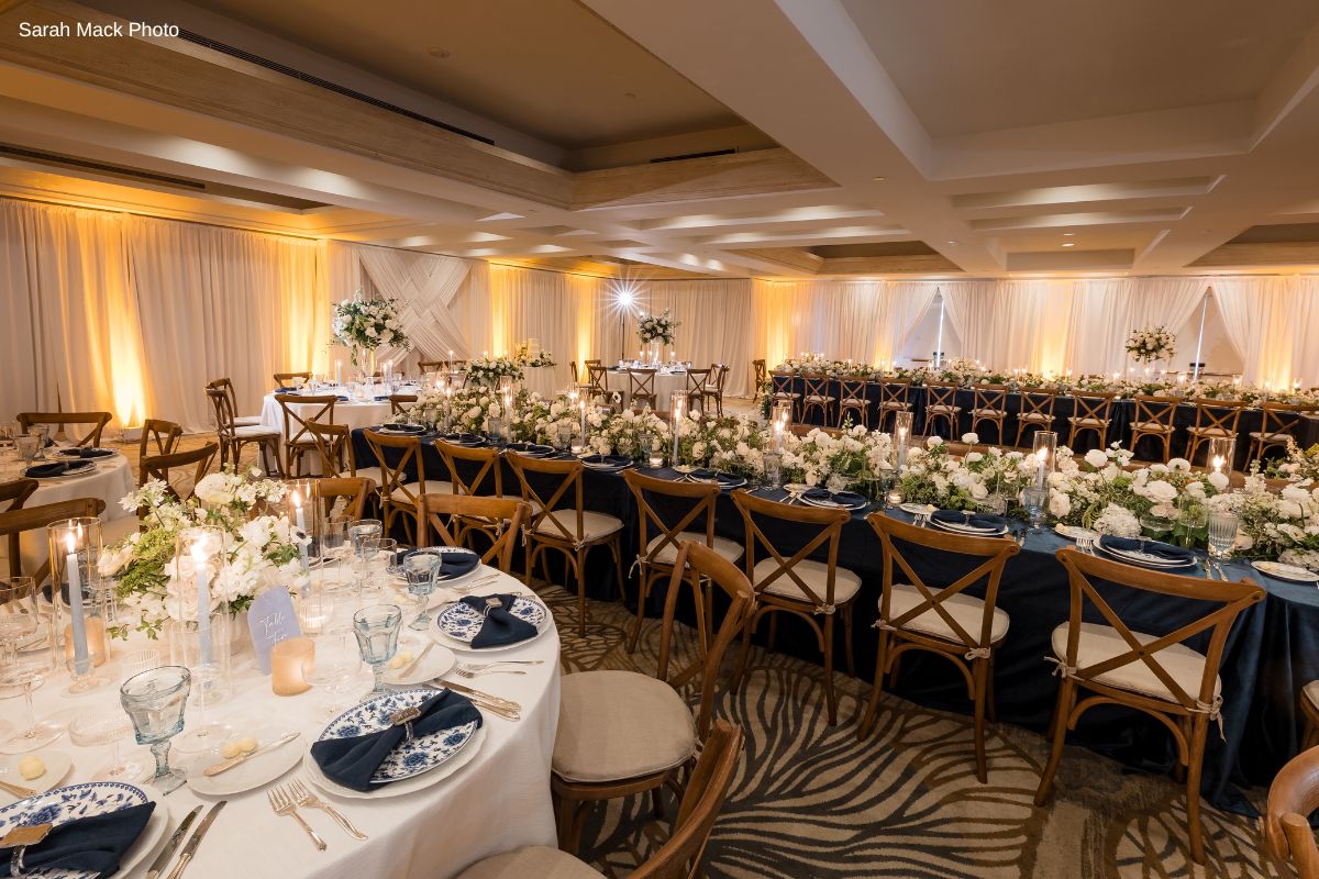 Elegant banquet hall set for an event, featuring long tables with floral centerpieces, wooden chairs, white tableware, and warm ambient lighting.