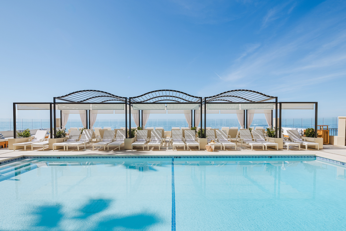 Outdoor swimming pool with clear blue water, lounge chairs, and canopy beds lined up along the poolside under a sunny sky. Ocean view is visible in the background.