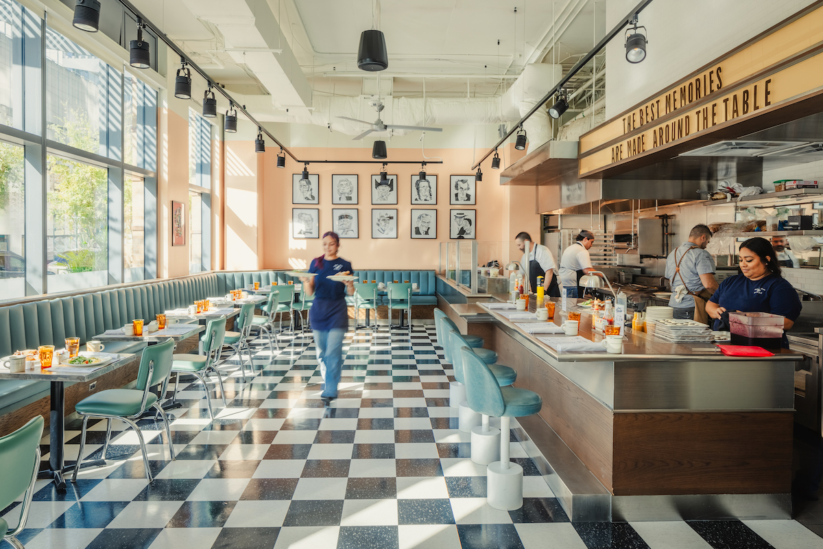 Bright diner with checkered floors, green booths, and a counter where staff prepare food; one person walks through the dining area.
