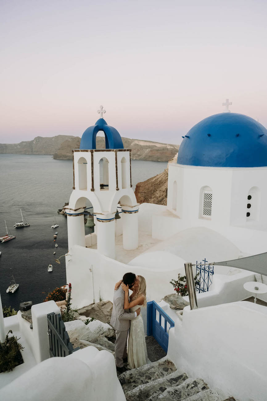 A couple embraces on white stone steps overlooking blue-domed buildings and the sea in Santorini at sunset.