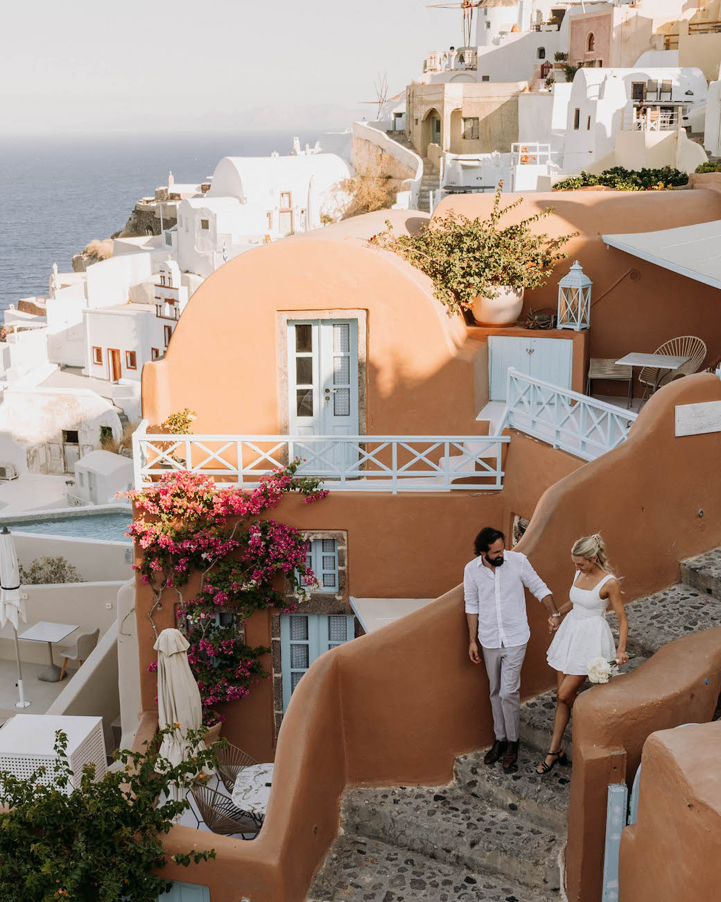 A couple walks down stone steps beside white and terracotta buildings with blue doors and pink flowers, overlooking the sea in a coastal Mediterranean town.