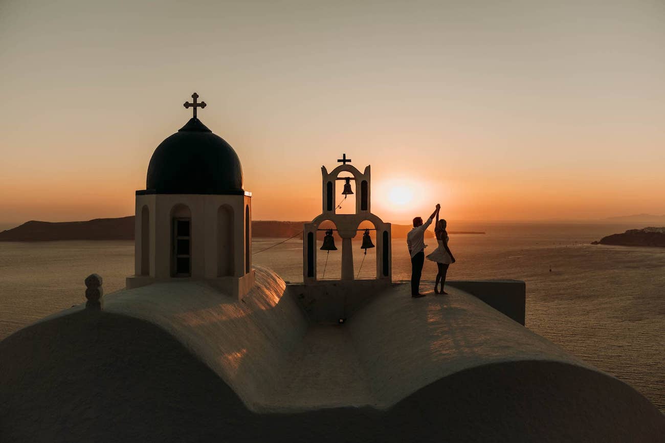 Two people stand on a white rooftop near a blue-domed church at sunset, overlooking the sea in Santorini, Greece.