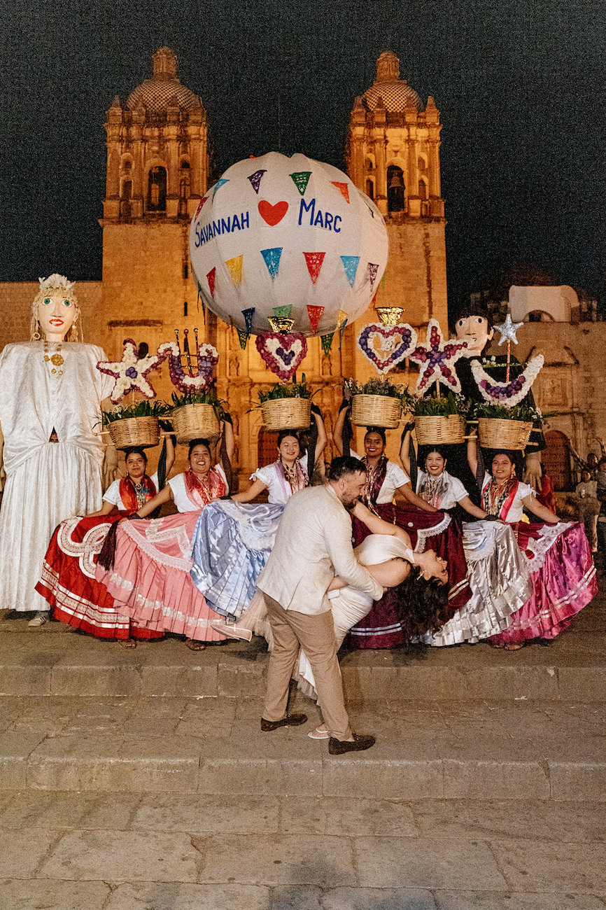 A couple dances in front of people in traditional Mexican dress with large baskets and puppets, in a plaza at night. A decorated balloon with names floats above.