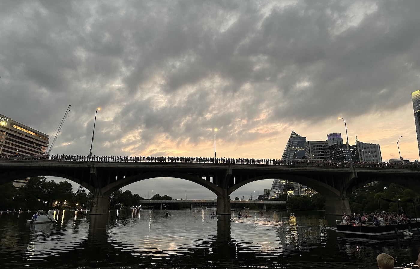A crowd stands along a bridge at dusk, with city buildings in the background and reflections on the water below.