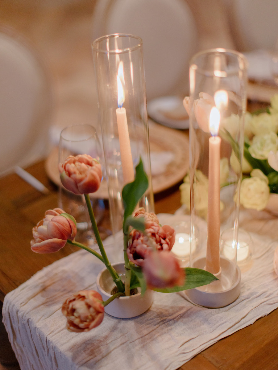 A wooden table set with a white runner, pink tulips in a vase, two lit taper candles in glass holders, and white plates.