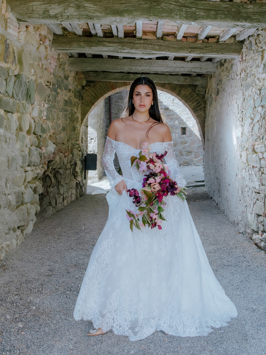 A woman in a white off-the-shoulder wedding dress holds a bouquet of flowers while standing in a stone archway.