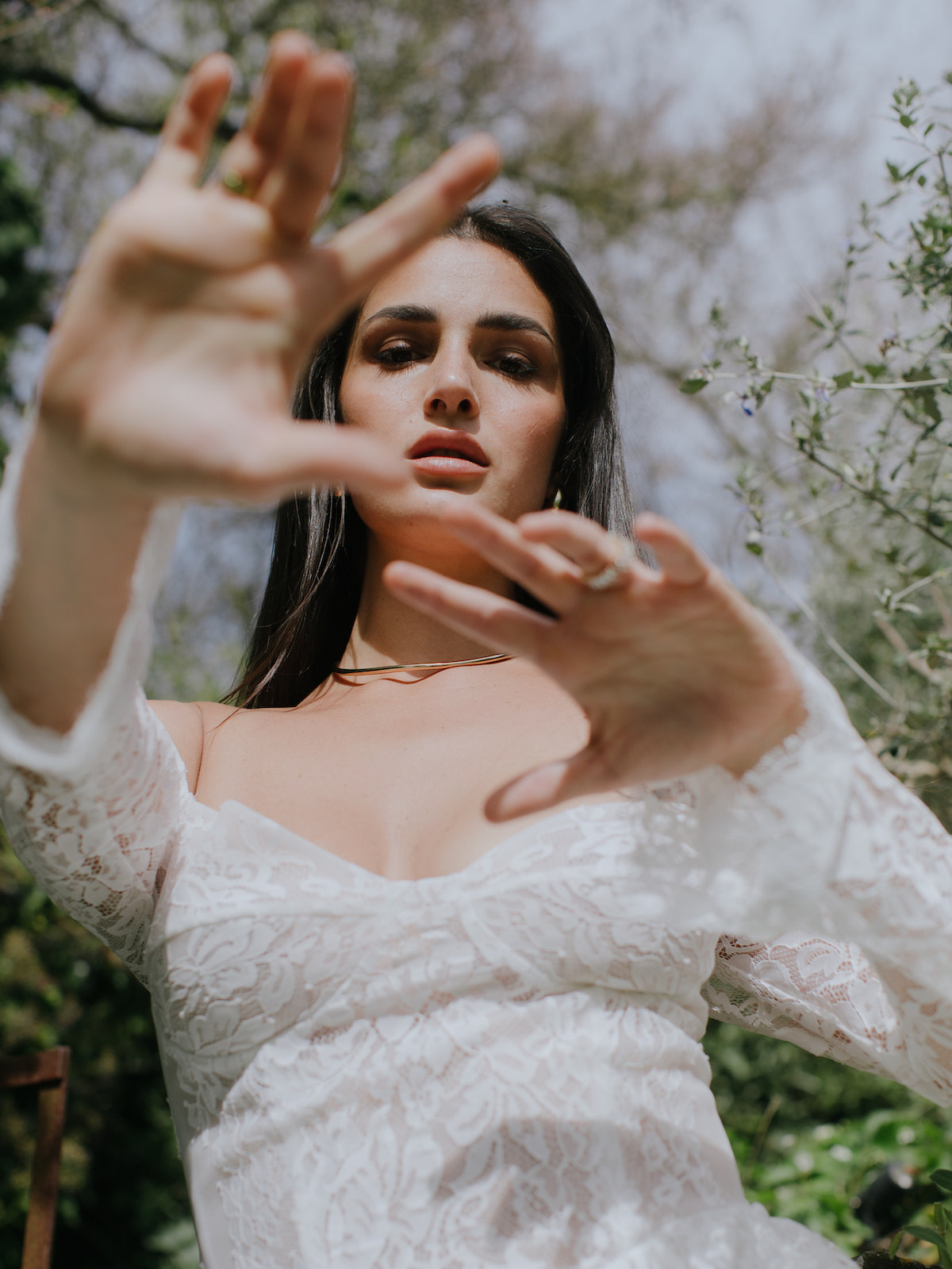 Woman in a white lace dress extends her hands toward the camera outdoors, with blurred greenery and trees in the background.