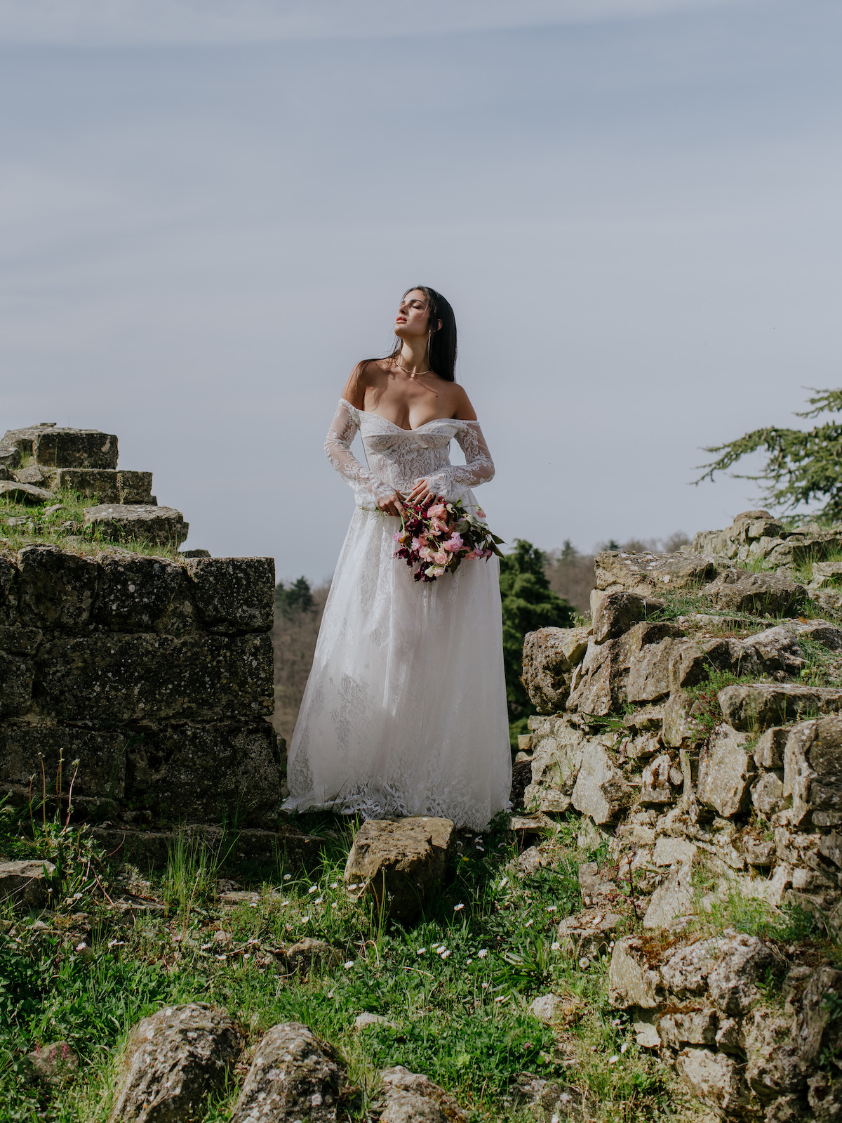 A woman in a white wedding dress stands among ancient stone ruins, holding a bouquet of flowers and looking upward.