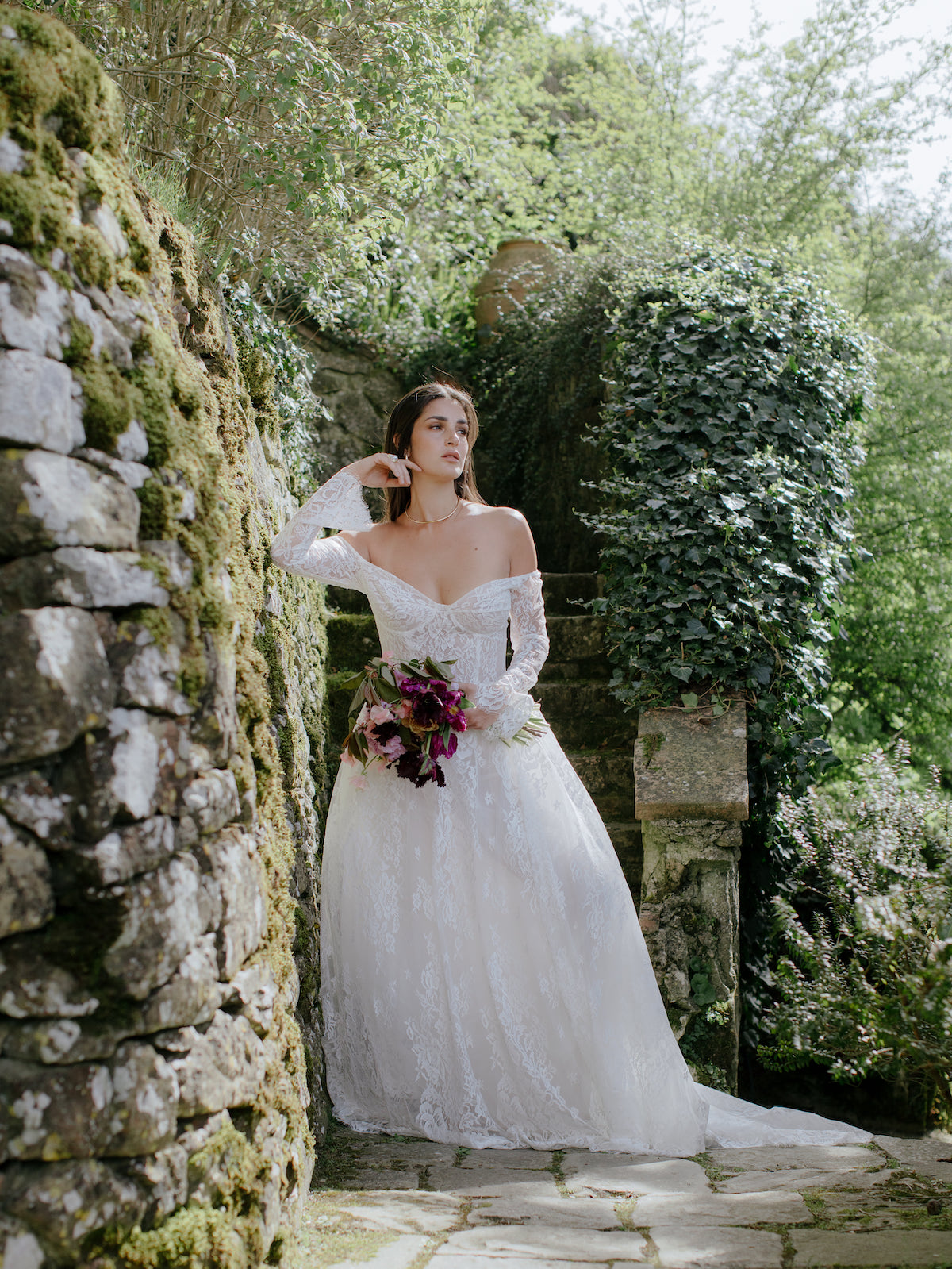 A woman in a white wedding dress holding a bouquet stands on a stone pathway surrounded by greenery and moss-covered walls.